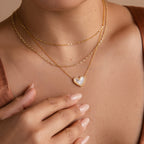Woman wearing layered gold necklaces, including the Pave Mother of Pearl Heart Necklace with a white pendant, and has manicured nails.