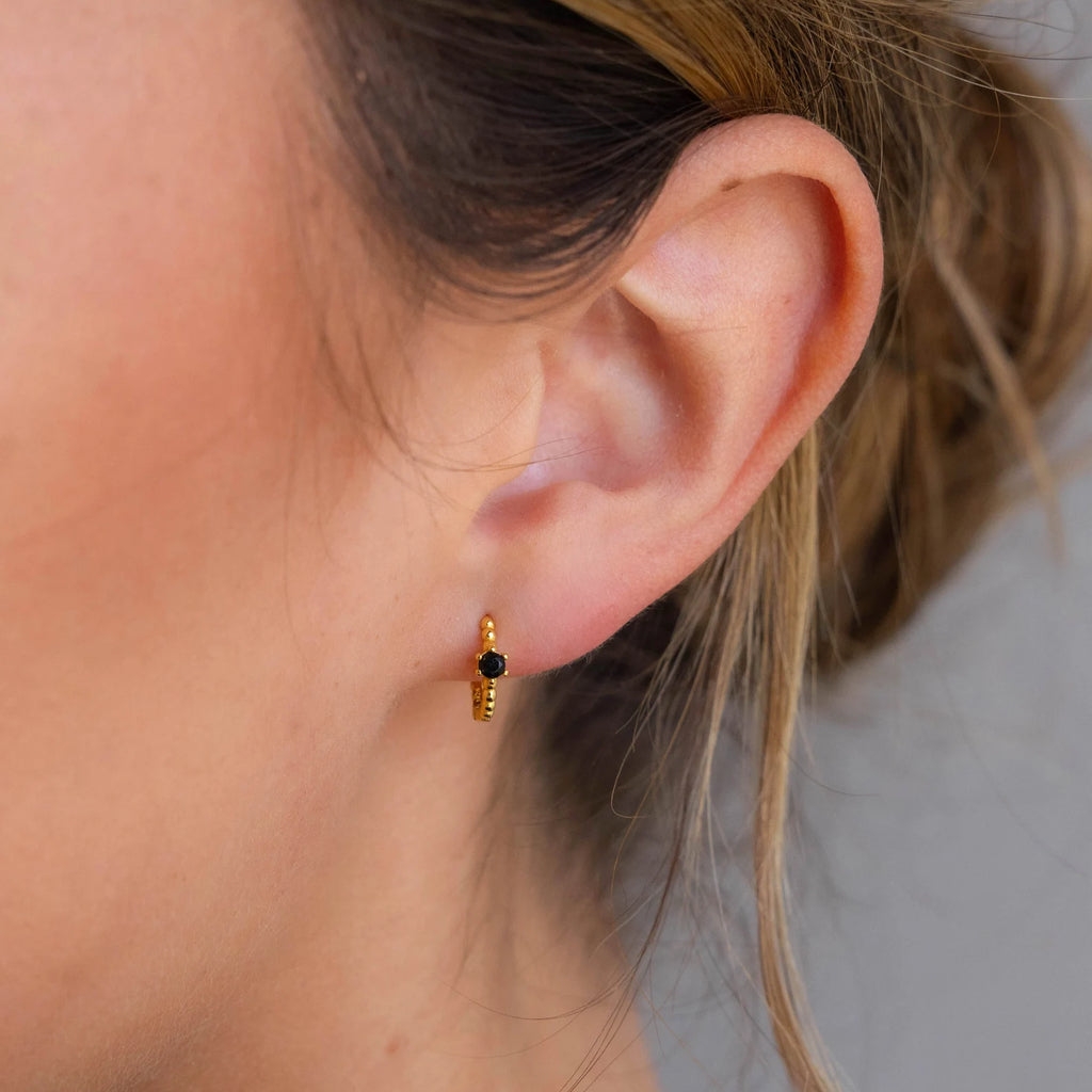 Close-up of a woman’s ear wearing the Onyx Beaded Huggies—a small gold hoop earring with a hexagonal Onyx charm.