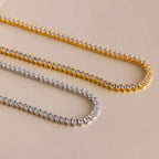 Two 3 Prong Tennis Necklaces—one gold, one silver—featuring round white stones and dainty chains are displayed on a beige surface for an elegant touch.