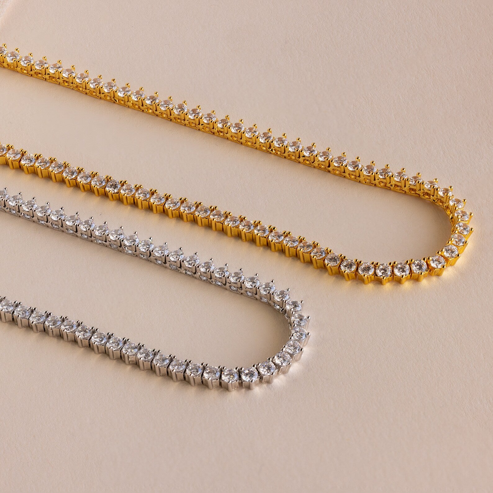 Two 3 Prong Tennis Necklaces—one gold, one silver—featuring round white stones and dainty chains are displayed on a beige surface for an elegant touch.
