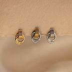 A close-up product shot of three individual Mini Triple Knot Earrings arranged in a horizontal row, suspended on the edge of a textured, natural-toned card. Each earring features three interlocking, high-polish bands forming a compact, elegant knot shape. The left earring is fully gold-toned, the center earring combines gold and silver tones for a mixed metal look, and the right earring is entirely silver-toned. 