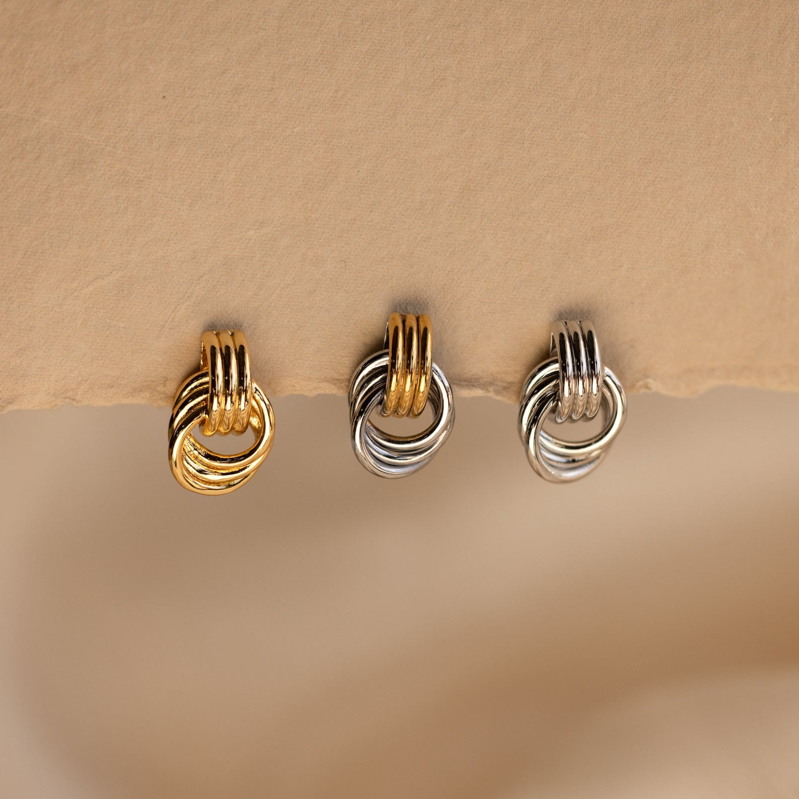 A close-up product shot of three individual Mini Triple Knot Earrings arranged in a horizontal row, suspended on the edge of a textured, natural-toned card. Each earring features three interlocking, high-polish bands forming a compact, elegant knot shape. The left earring is fully gold-toned, the center earring combines gold and silver tones for a mixed metal look, and the right earring is entirely silver-toned. 