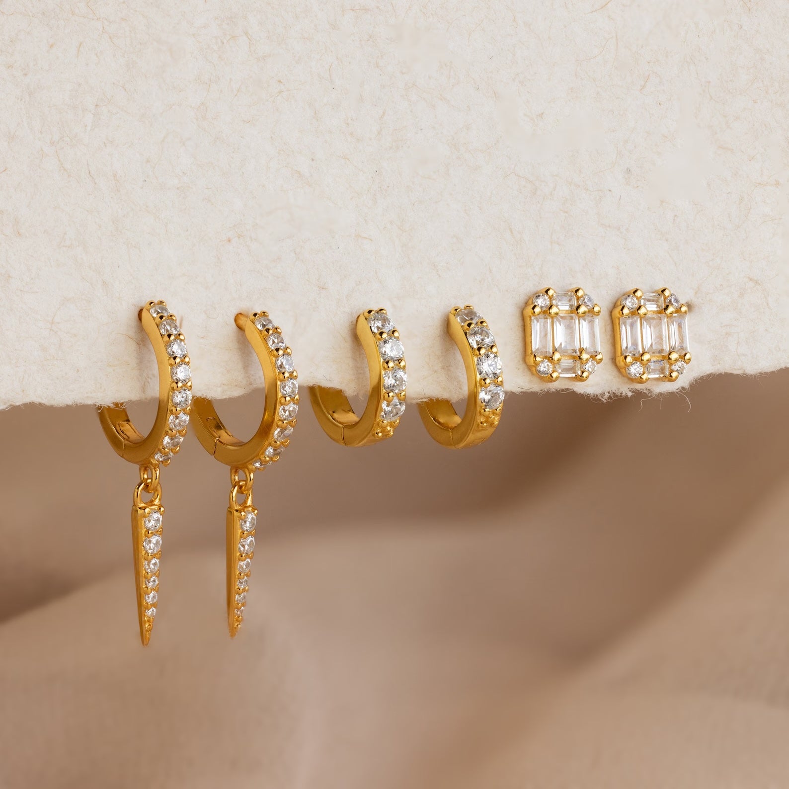 Three pairs of gold earrings with small clear stones rest on a soft, beige surface—ideal for adding to your Mystery Earrings Box.