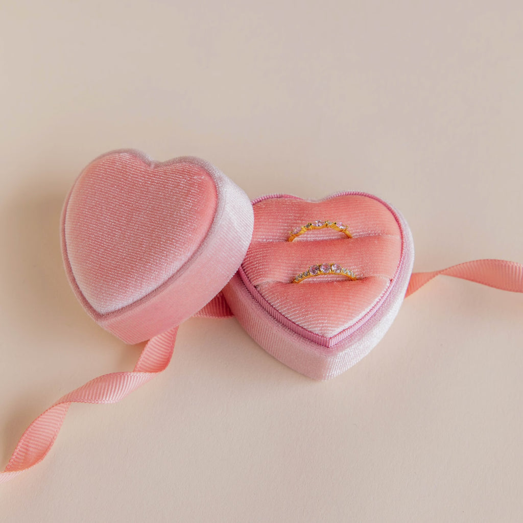 Two gold rings with diamonds are nestled in a Pink Velvet Heart Ring Box, adorned with a matching ribbon for a chic, coquette style touch, set against a light background.