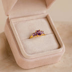 A Pink Suede Ring Box beautifully displays a gold ring with three purple gemstones, featuring a minimalist design.