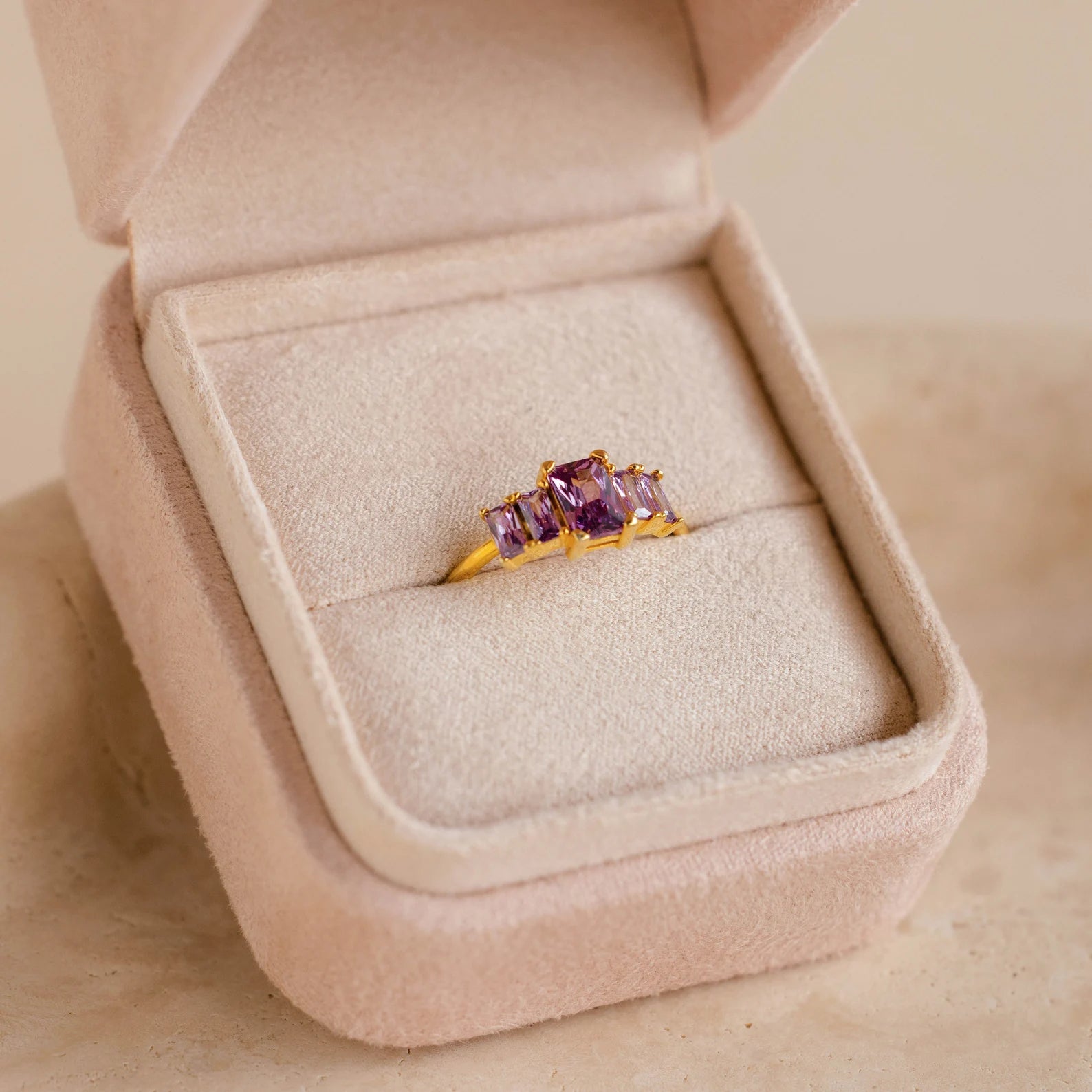A Pink Suede Ring Box beautifully displays a gold ring with three purple gemstones, featuring a minimalist design.