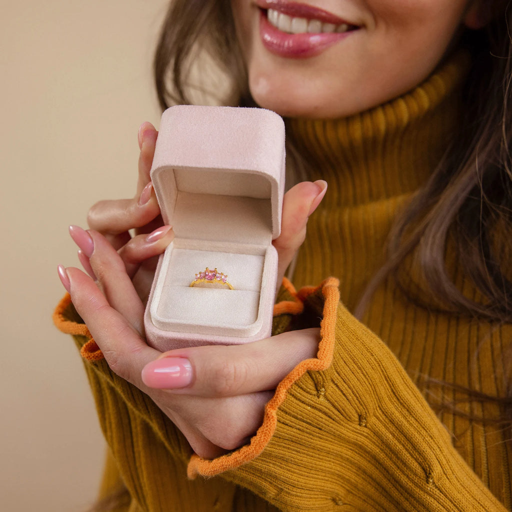 A woman in a mustard sweater smiles while holding an open Pink Suede Ring Box with a jeweled ring inside.