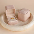 A Pink Suede Ring Box sits open, displaying a gold ring with purple stones next to a closed jewelry box, both atop a round stone dish for a chic, minimalist look.