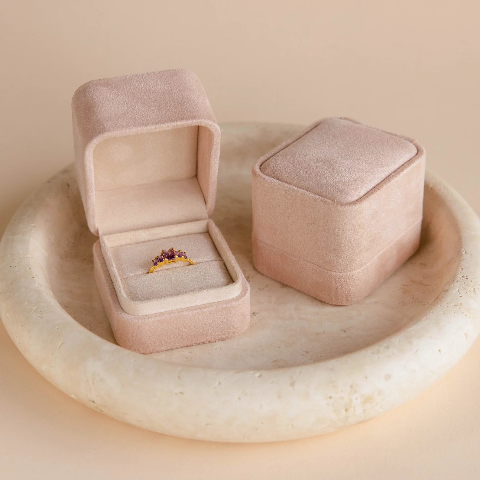 A Pink Suede Ring Box sits open, displaying a gold ring with purple stones next to a closed jewelry box, both atop a round stone dish for a chic, minimalist look.