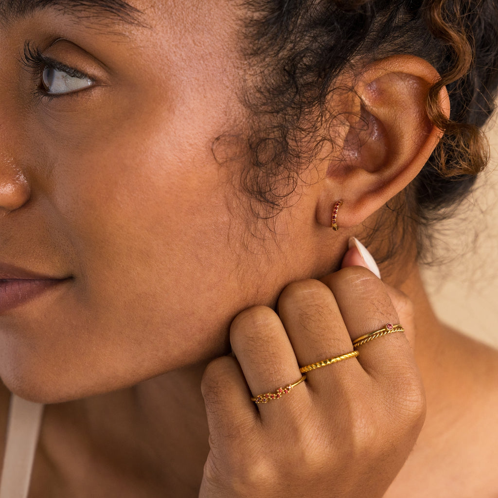 Close-up of a woman wearing gold rings and Dainty Pave Birthstone Huggies in her ear.