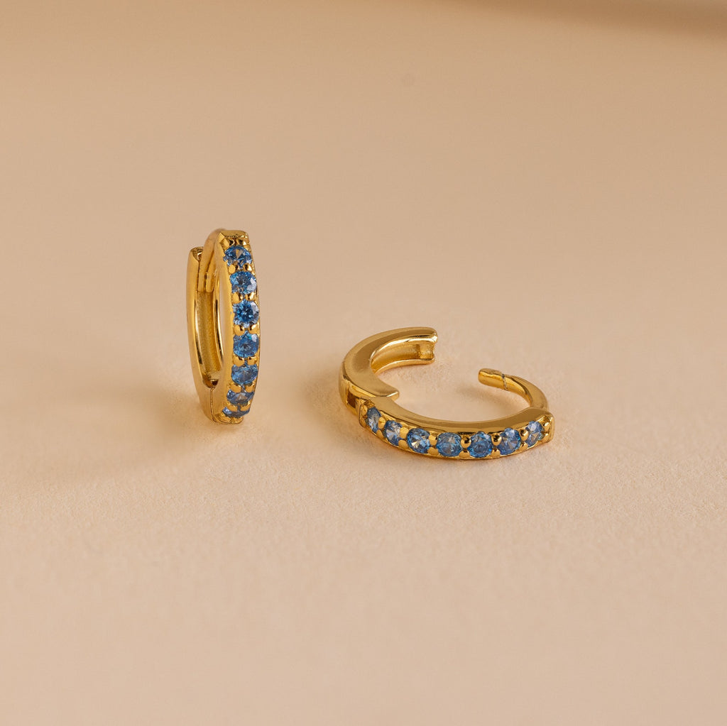The Dainty Pave Birthstone Huggies are small gold hoop earrings with blue gemstones, displayed on a beige surface—perfect for adding everyday sparkle.