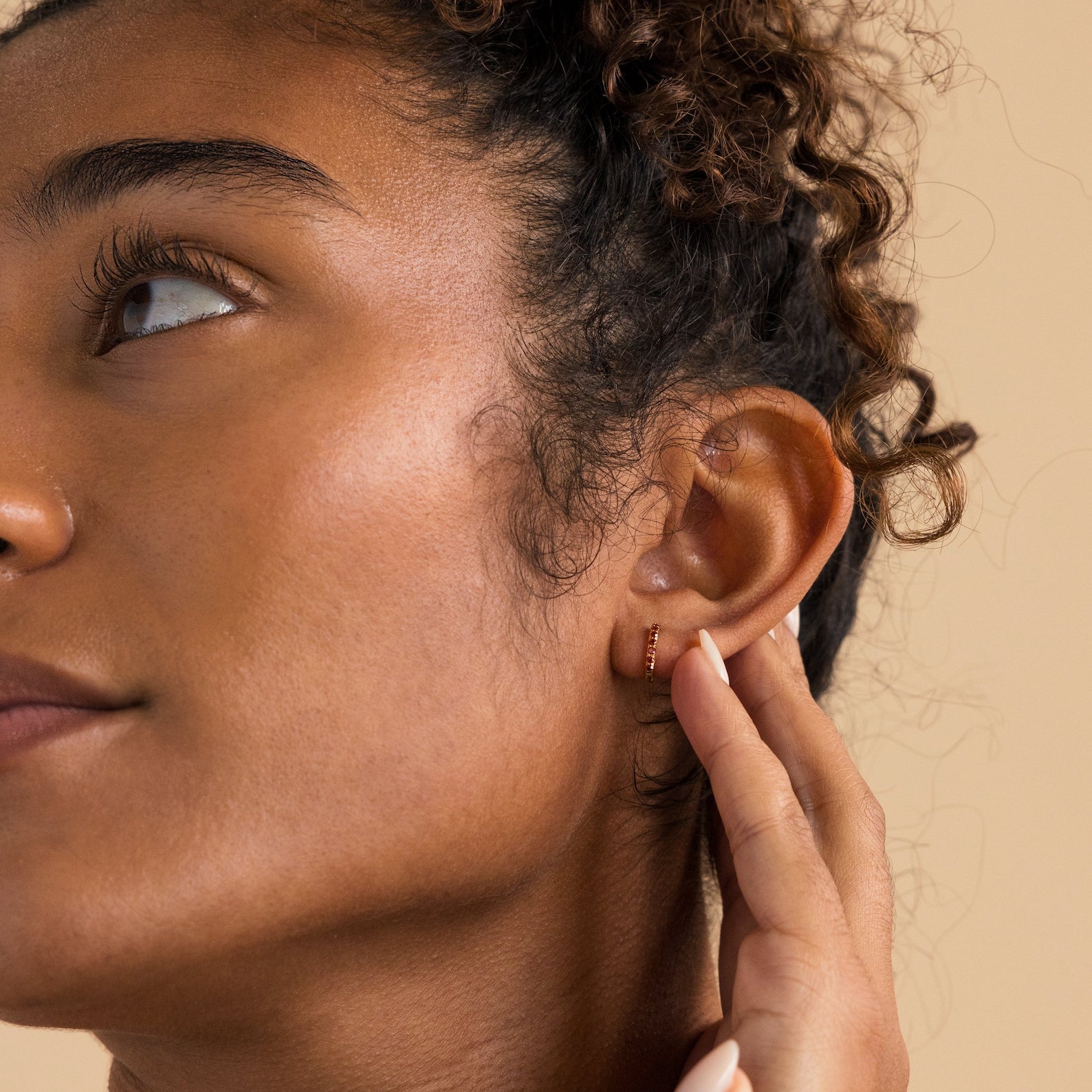 A woman with curly hair touches her ear, displaying the Dainty Pave Birthstone Huggies—small gold hoop earrings accented with sparkling gemstones—against a neutral background.