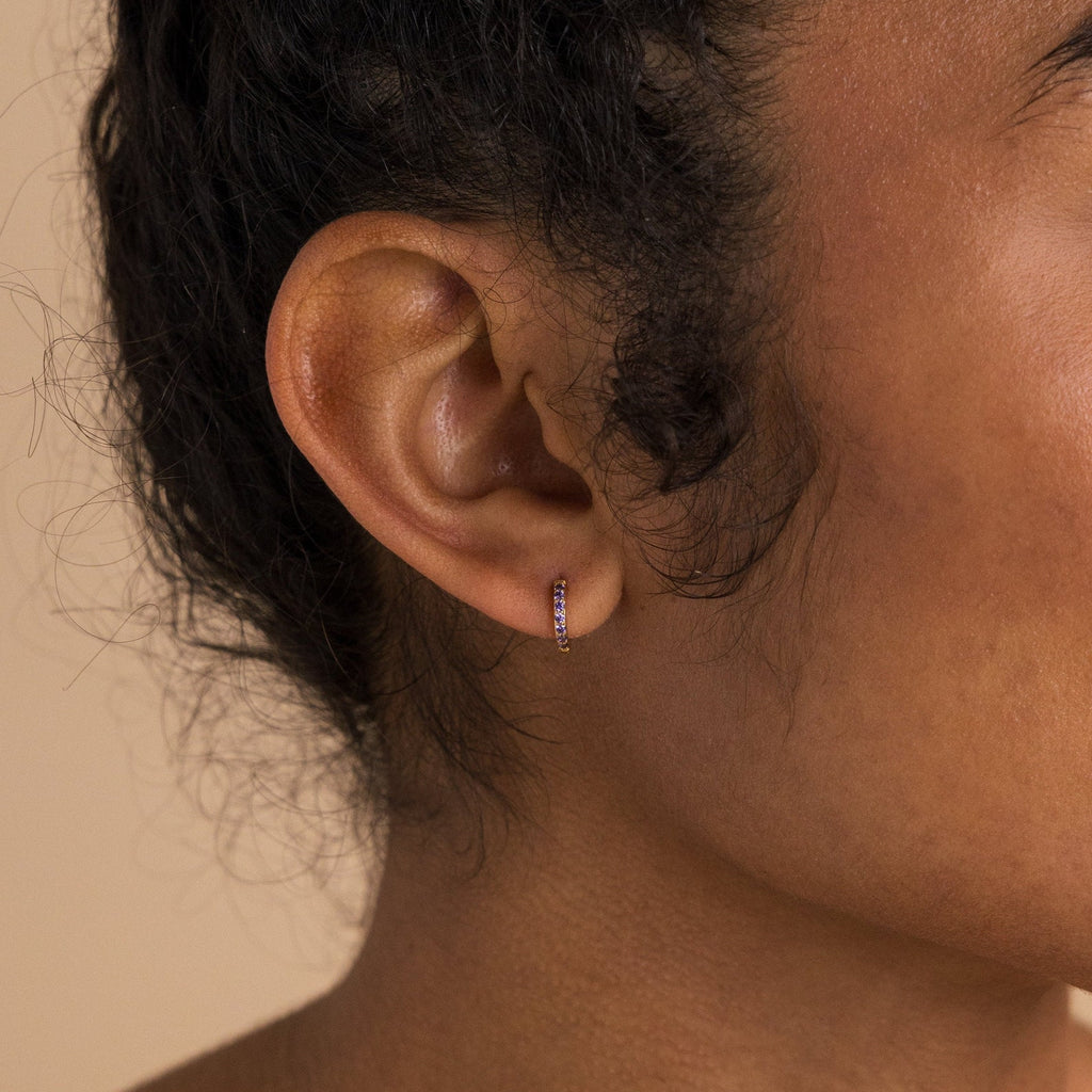 Close-up of a woman's ear wearing Dainty Pave Birthstone Huggies—small hoop earrings with purple gemstones—against a neutral background.