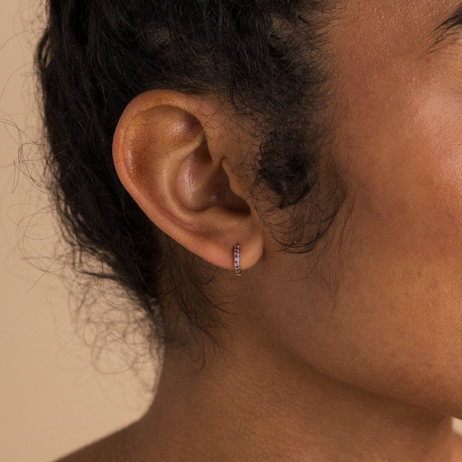 Close-up of a woman's ear wearing Dainty Pave Birthstone Huggies—small hoop earrings with purple gemstones—against a neutral background.