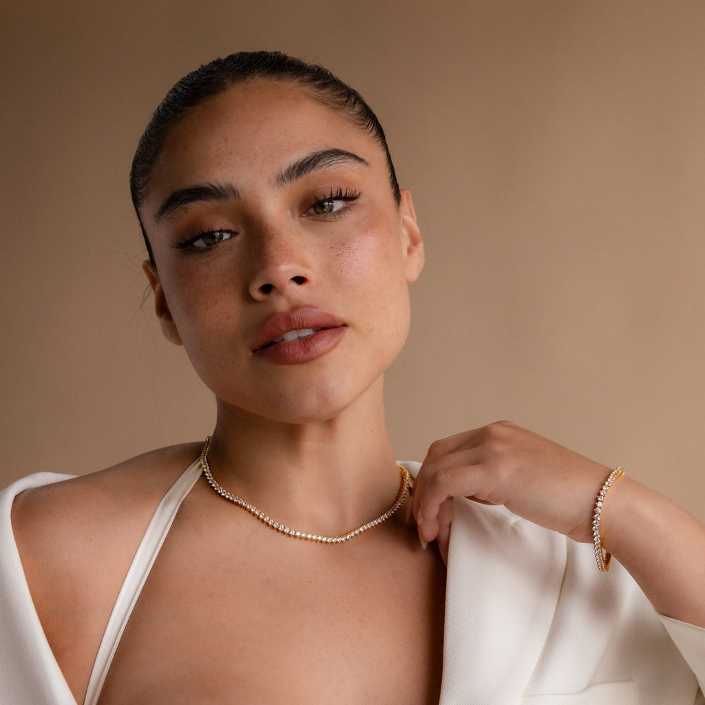 A woman with slicked-back hair wears a white outfit, gold bracelet, and the 3 Prong Tennis Necklace featuring diamond gemstones against a beige background.