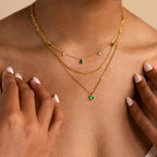 A person wears a layered gold necklace with green and white gems, hands near their collarbone—ideal as an everyday accessory or styled with the Skinny Mariner Chain Necklace for extra flair.