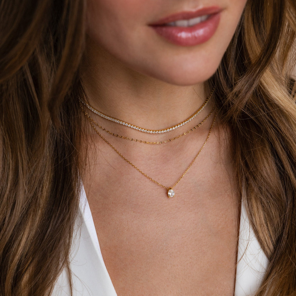 Layered necklace set featuring a gold choker with clear stones, a delicate gold chain, and a longer gold chain with a single Pear-cut crystal pendant, worn with a white V-neck top.