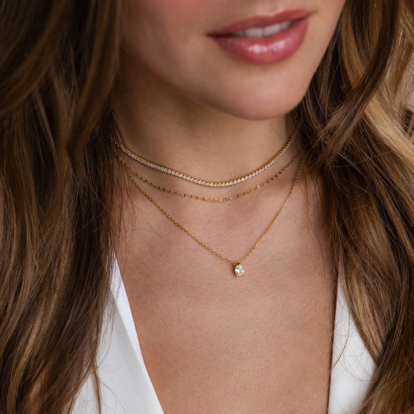 Layered necklace set featuring a gold choker with clear stones, a delicate gold chain, and a longer gold chain with a single Pear-cut crystal pendant, worn with a white V-neck top.