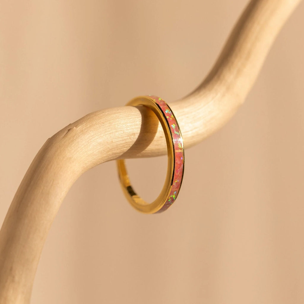 A gold ring from our Opal Inlay Rings Set, featuring a pink opal inlay, rests on a curved light wood branch against a beige background—capturing the graceful elegance of a Pink Flamingo.