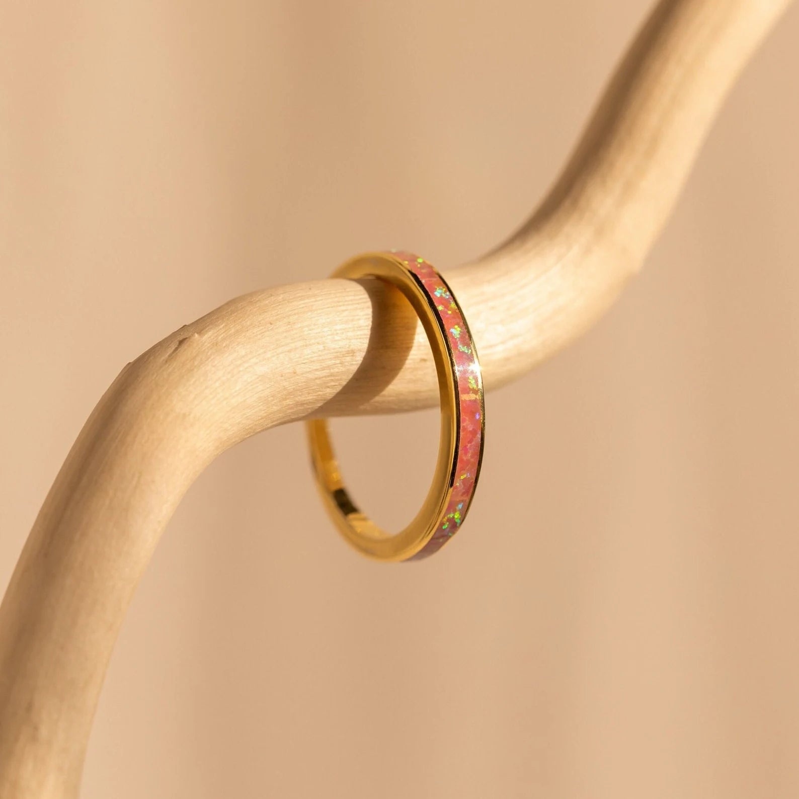 A gold ring from our Opal Inlay Rings Set, featuring a pink opal inlay, rests on a curved light wood branch against a beige background—capturing the graceful elegance of a Pink Flamingo.