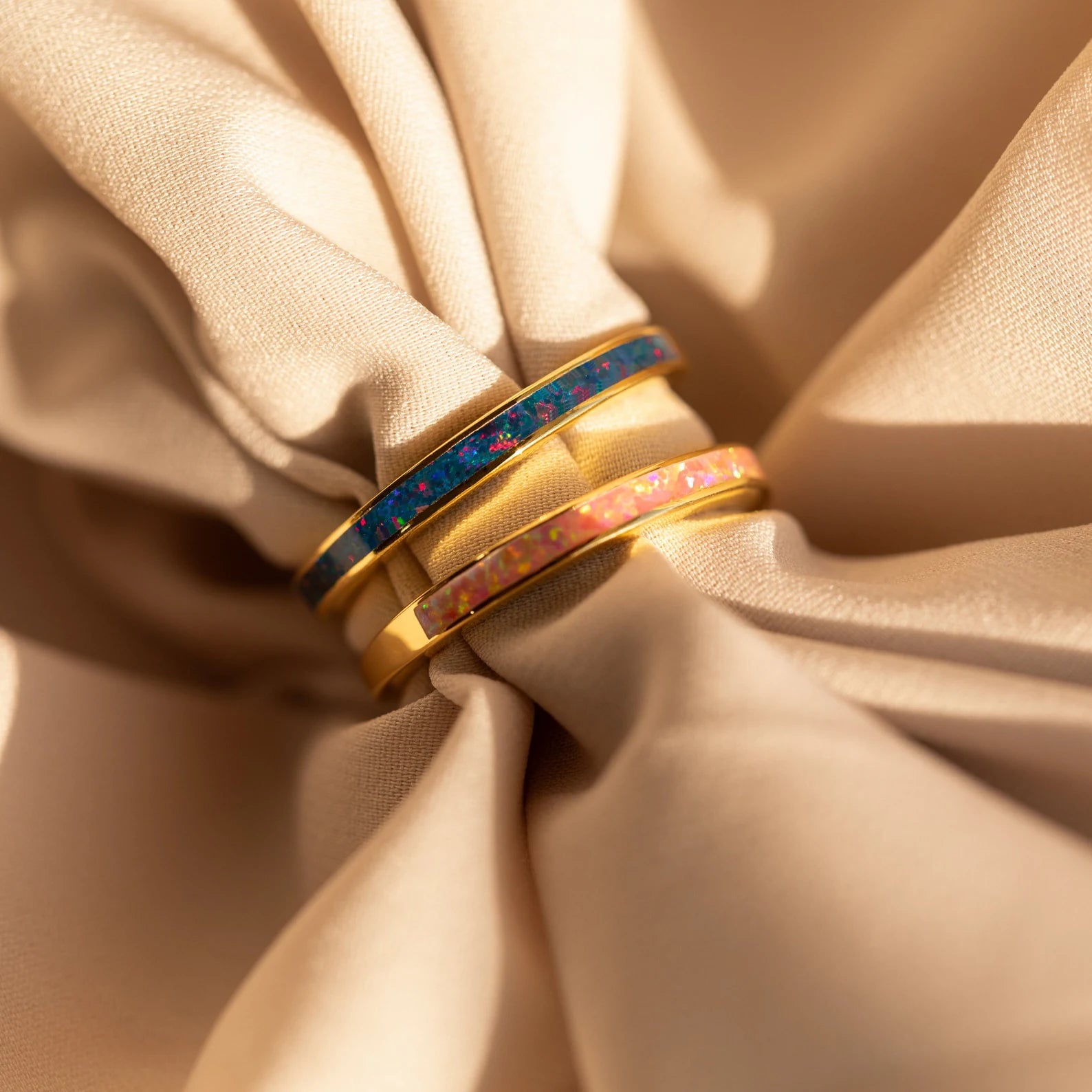 The Opal Inlay Rings Set, featuring gold bands with indigo blue accents, rests on beige satin fabric under soft, warm lighting.