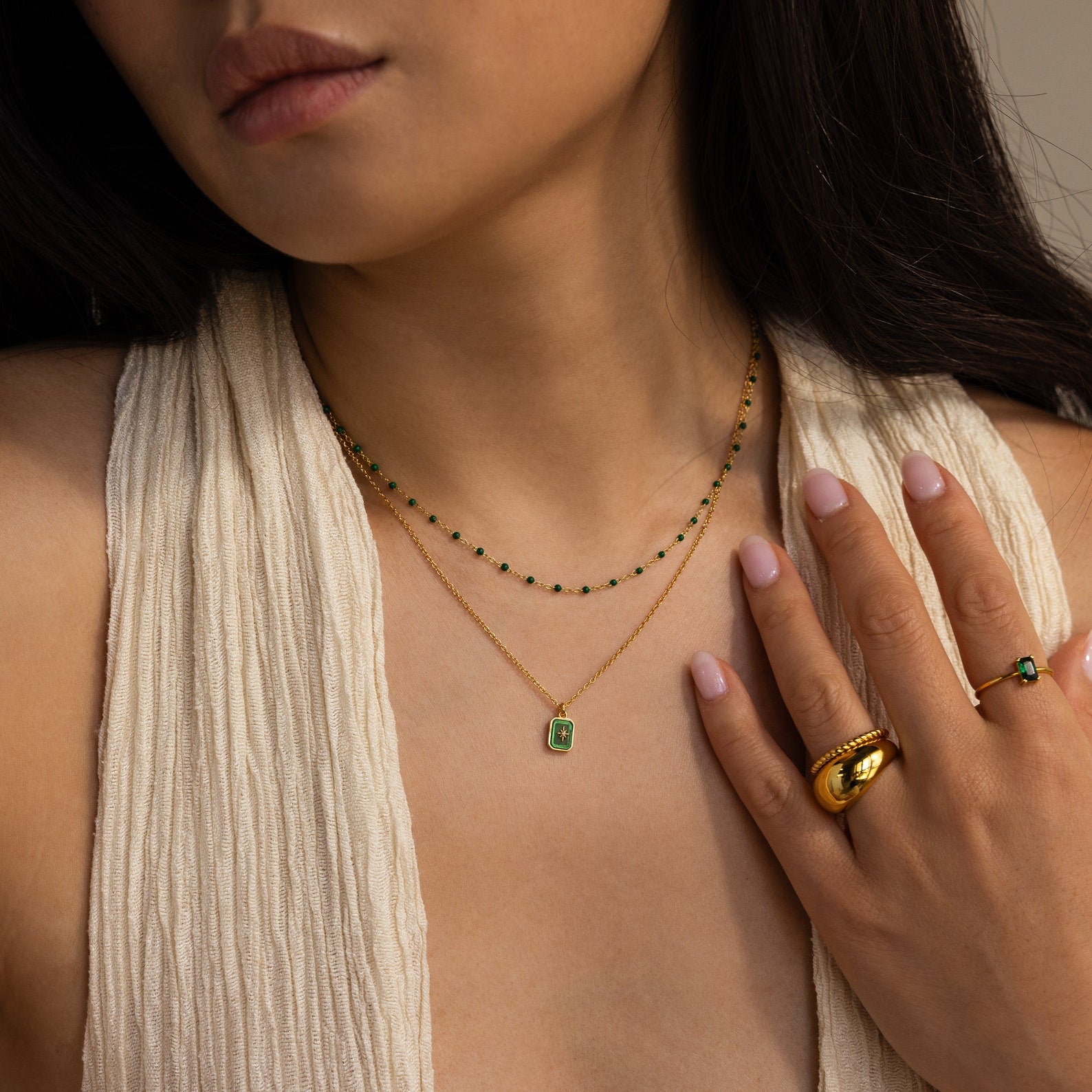 A woman wears layered gold necklaces, featuring the North Star Birthstone Necklace with green stones, a gold ring, and a sleeveless cream top.