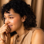 A smiling woman with curly hair and a nose ring wears a beige top and Birthstone Flower Studs earrings while sitting in front of beige curtains.