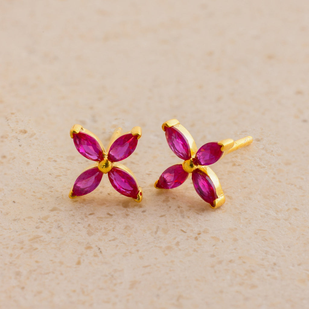 The Birthstone Flower Studs feature four marquise pink stones in a gold floral design, offering an elegant touch to any look. Perfect for everyday wear, these gemstone earrings are displayed on a beige surface.