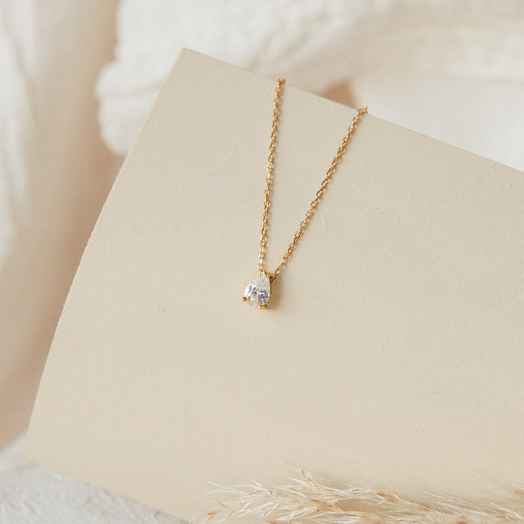 The Dainty Diamond Necklace Set features a gold chain with a single teardrop-shaped clear gemstone pendant, elegantly displayed on a light beige curved surface.