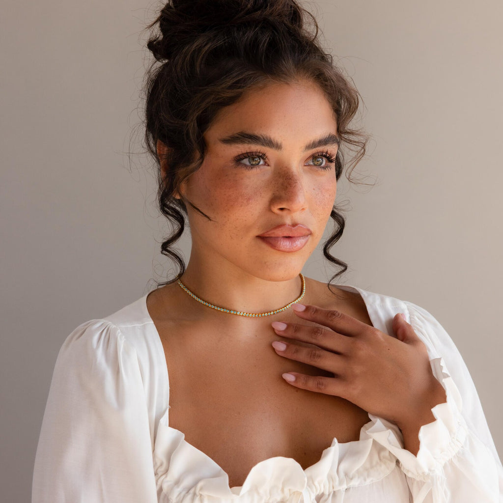 A woman with curly hair in a bun, wearing a white top and the Turquoise Bezel Tennis Necklace, touches her chest while gazing off to the side.