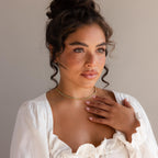A woman with curly hair in a bun, wearing a white top and the Turquoise Bezel Tennis Necklace, touches her chest while gazing off to the side.