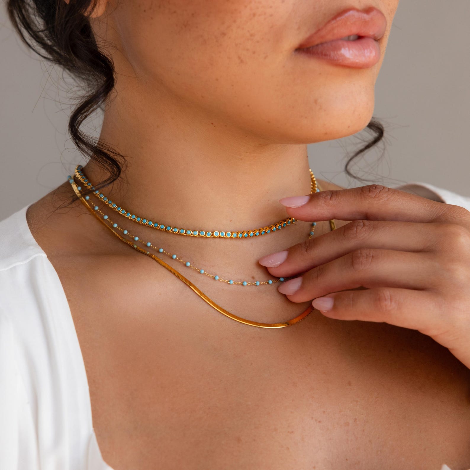 A woman in a white top with manicured nails touches her neck, showcasing the Turquoise Bezel Tennis Necklace layered with a gold choker.