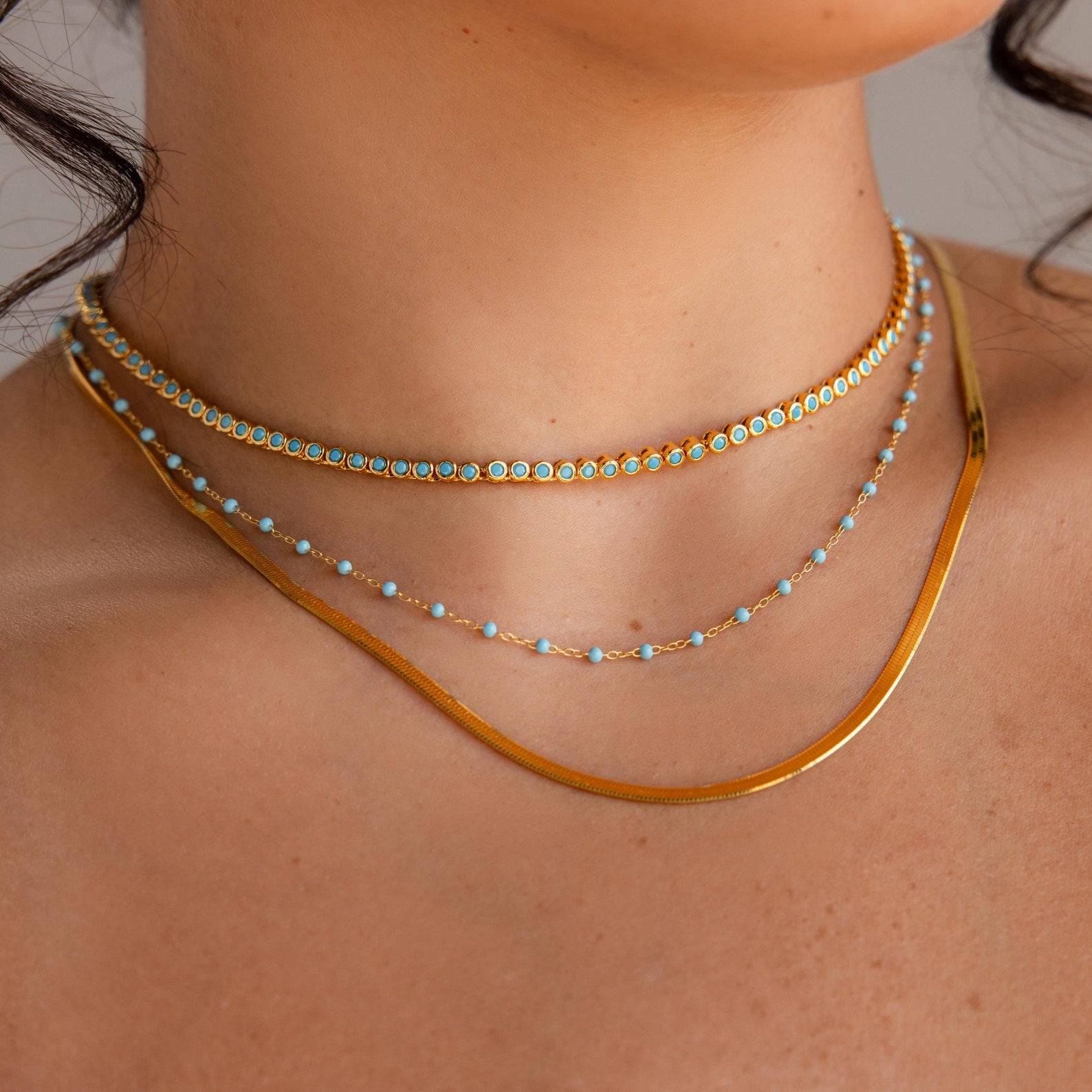Close-up of a woman's neck adorned with the Turquoise Bezel Tennis Necklace, layered with gold necklaces and a delicate choker for a chic, elegant look.