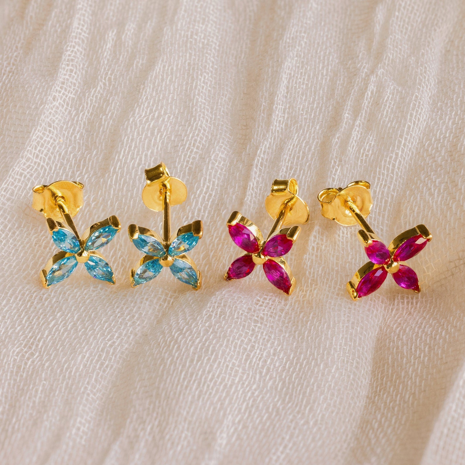 Two pairs of Birthstone Flower Studs, featuring blue and pink marquise-cut stones in gold, rest on cream fabric—ideal for adding color to your collection or wearing as personalized earrings.