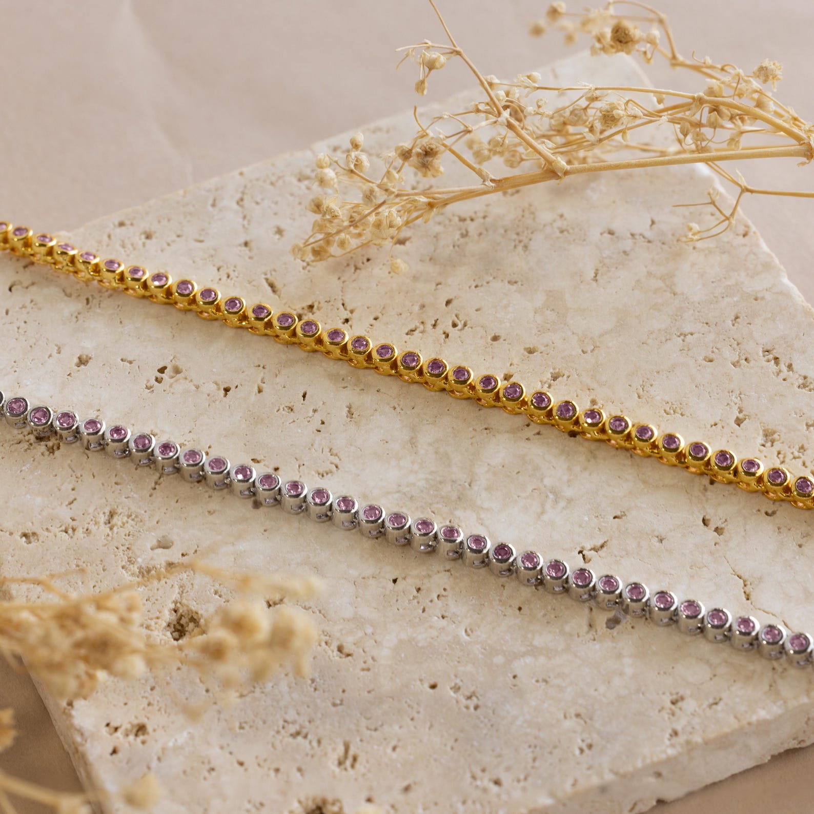 Gold and silver tennis bracelets with pink gemstones rest on a beige stone slab, accented by dried flowers—an ideal match with our Pink Bezel Tennis Necklace.
