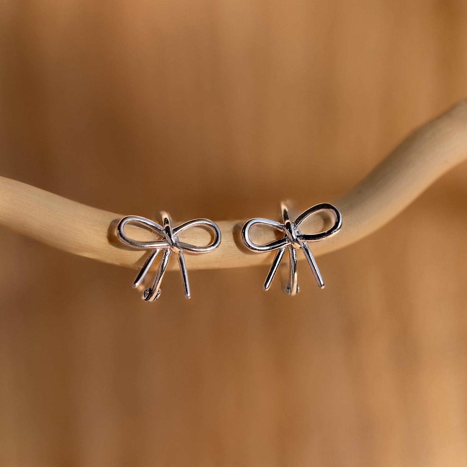 Classic Ribbon Bow Huggies gracefully dangle from a light branch with a soft brown backdrop—the perfect coquette earrings for adding a feminine touch.