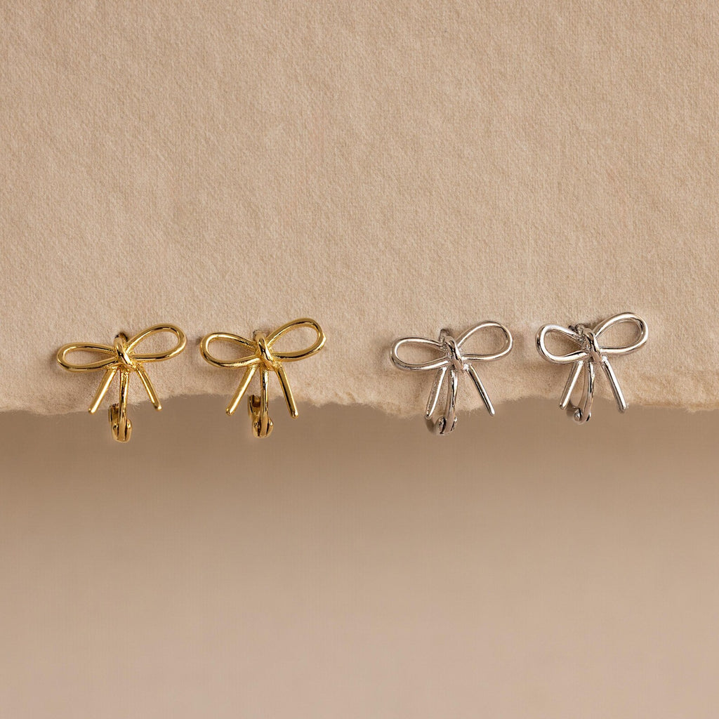 Two pairs of Classic Ribbon Bow Huggies in gold and silver are displayed on a textured beige surface, adding a touch of feminine elegance.