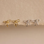 Two pairs of Classic Ribbon Bow Huggies in gold and silver are displayed on a textured beige surface, adding a touch of feminine elegance.