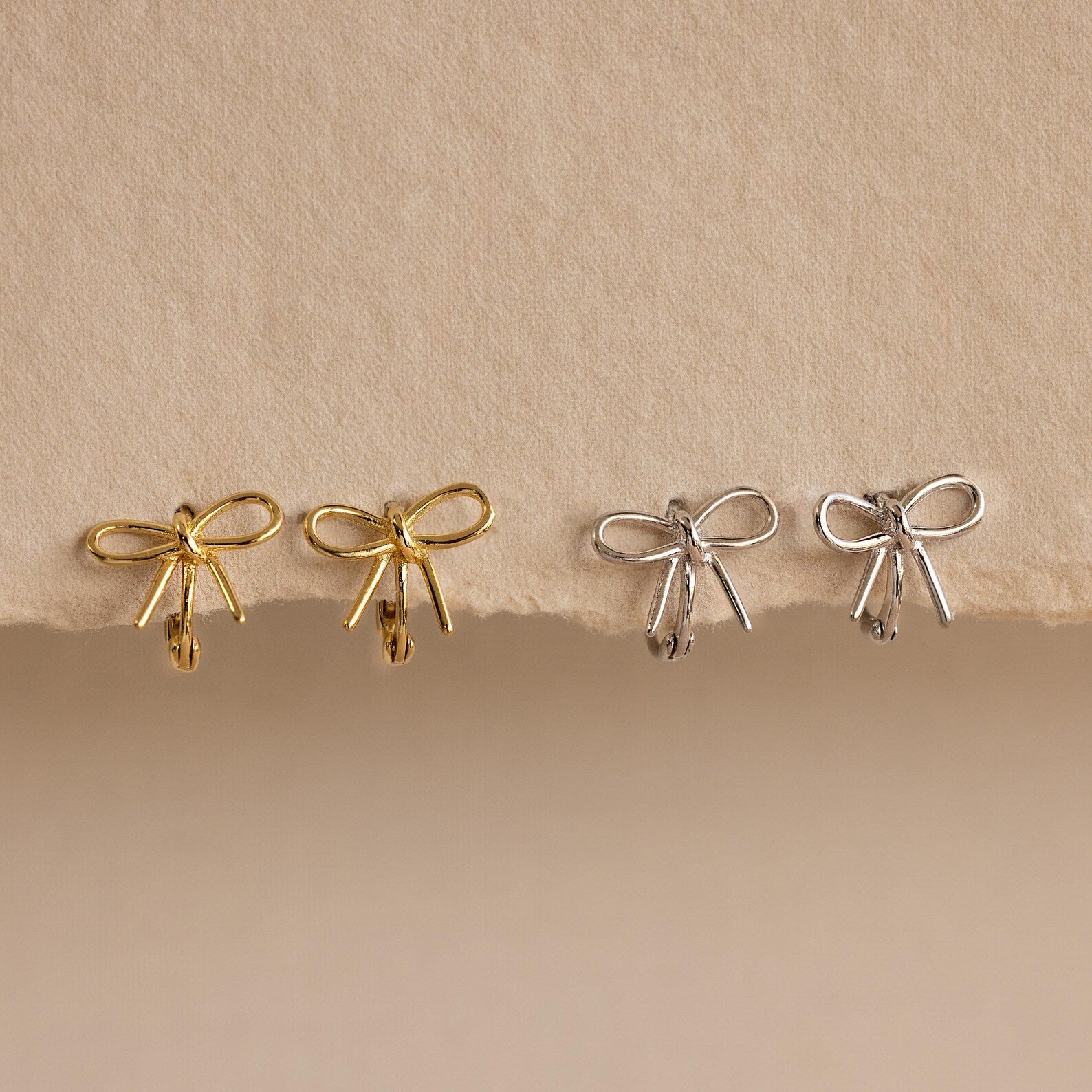 Two pairs of Classic Ribbon Bow Huggies in gold and silver are displayed on a textured beige surface, adding a touch of feminine elegance.