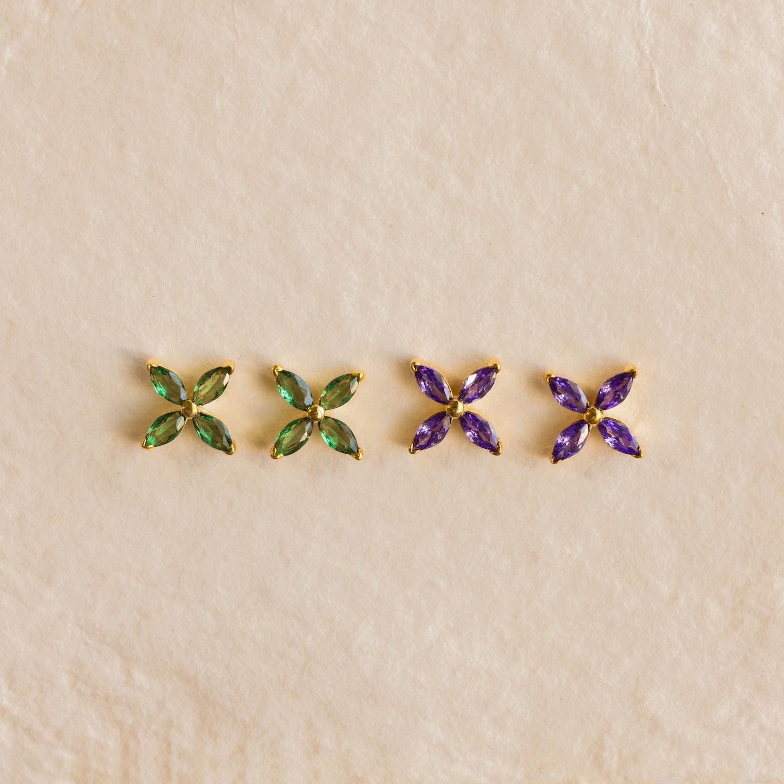 Two pairs of Birthstone Flower Studs—one with green and one with purple gemstones—are showcased on a beige background, making them perfect personalized earrings for any occasion.