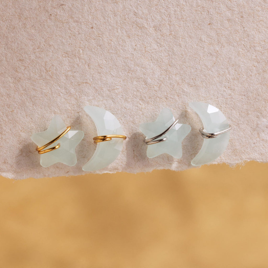 Four Aquamarine Moon & Star Studs, featuring translucent star and moon designs with gold and silver accents, rest delicately on textured paper.