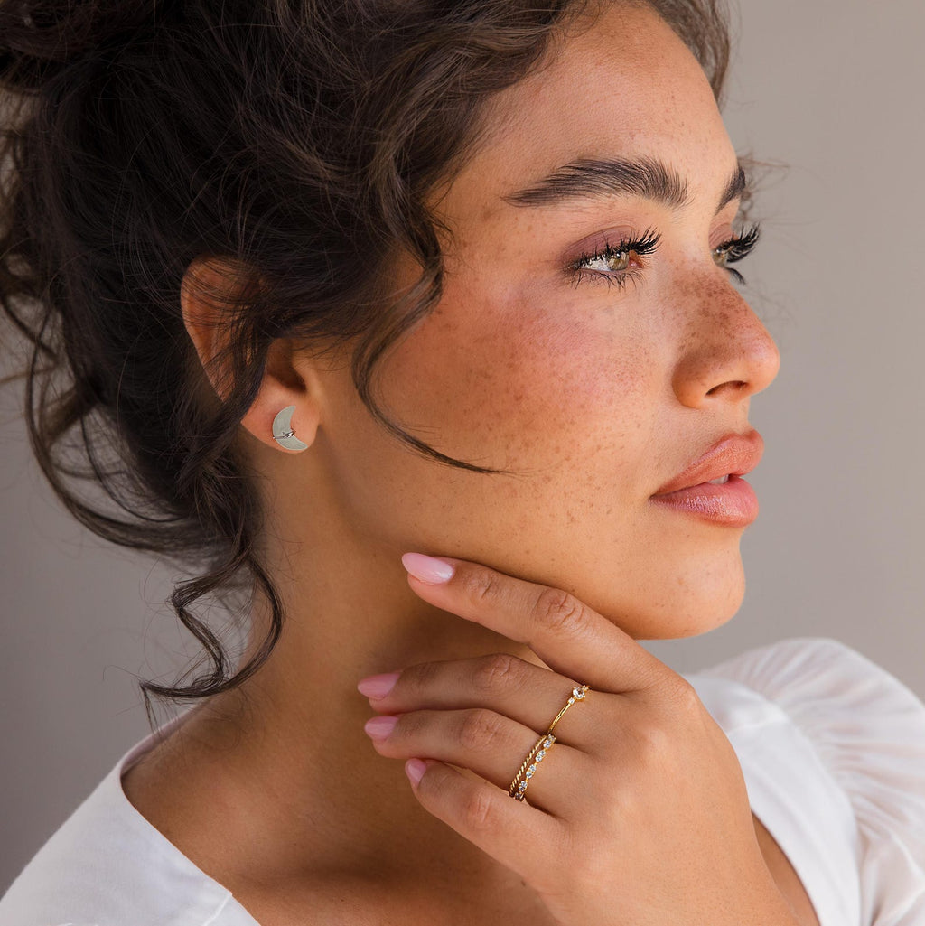 Woman with curly hair wearing Aquamarine Moon & Star Studs—elegant celestial earrings—paired with gold rings, touching her chin and gazing to the side.