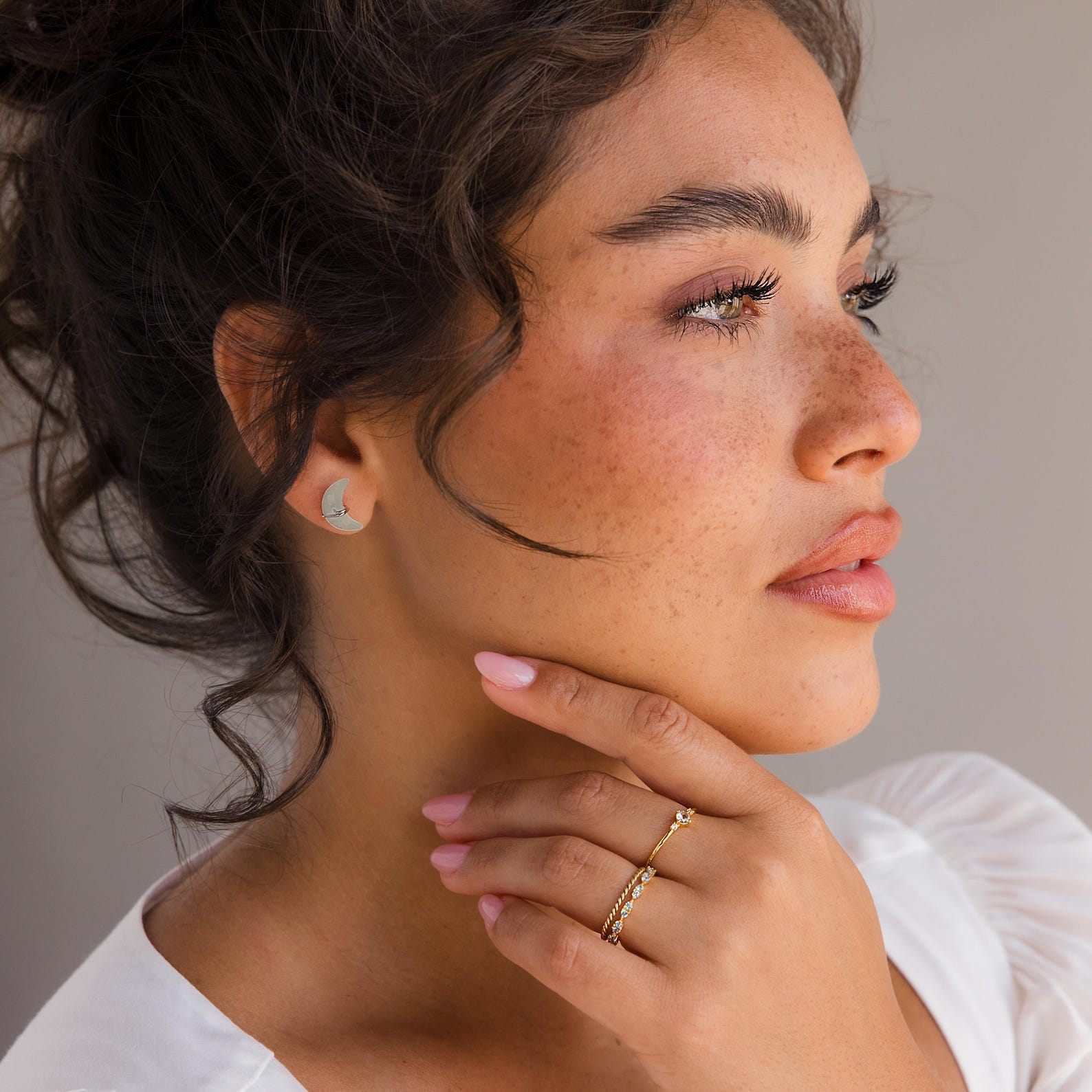 Woman with curly hair wearing Aquamarine Moon & Star Studs—elegant celestial earrings—paired with gold rings, touching her chin and gazing to the side.
