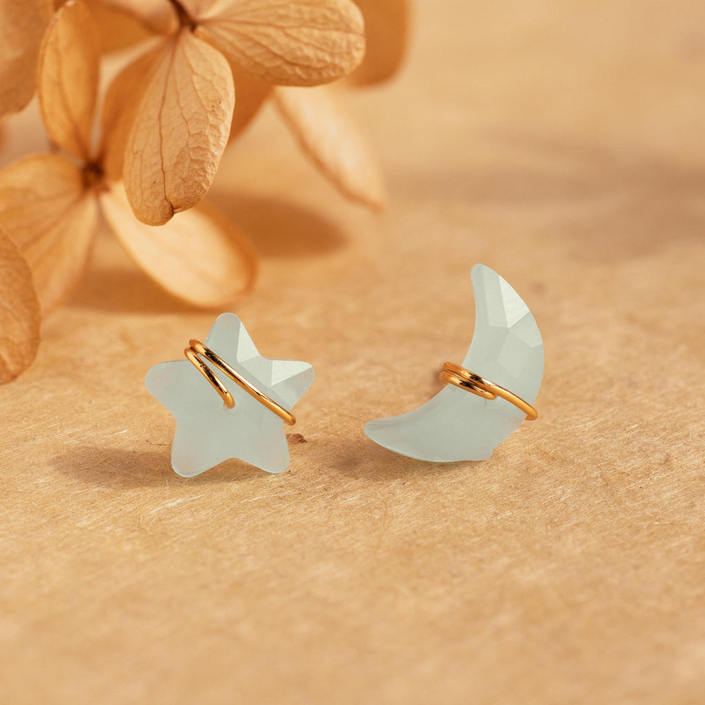 Aquamarine Moon & Star Studs featuring pale blue celestial shapes with gold accents are displayed on a beige surface with dried flowers in the background.