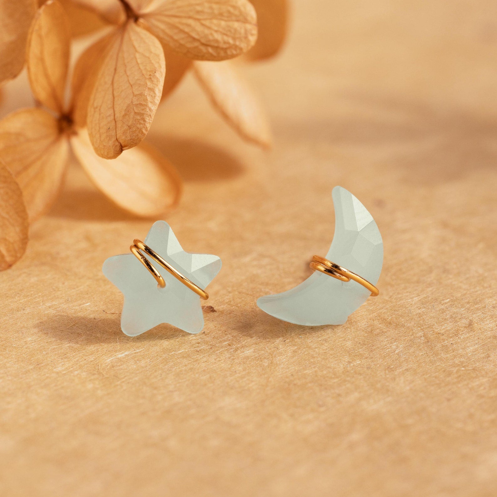 Aquamarine Moon & Star Studs featuring pale blue celestial shapes with gold accents are displayed on a beige surface with dried flowers in the background.