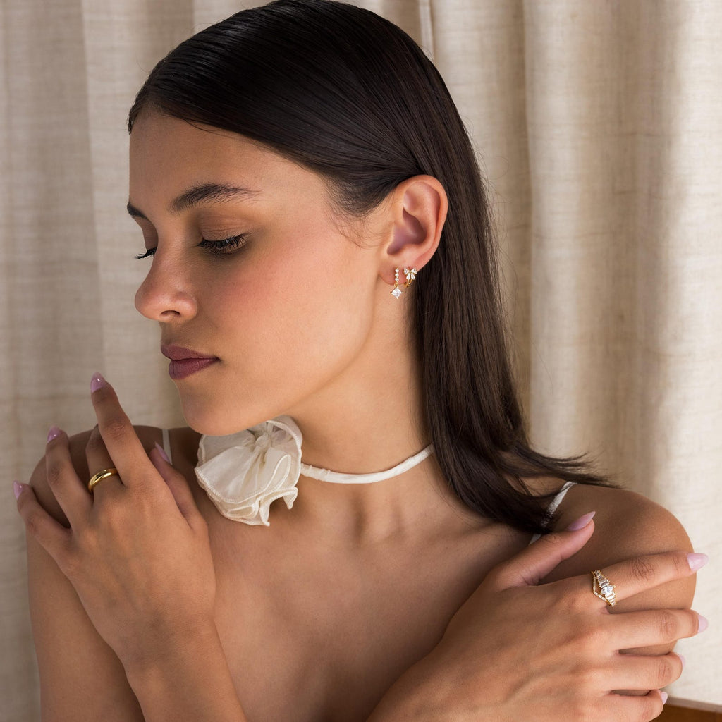 Model showcases Diamond Ribbon Bow Huggies paired with Diamond Drop Earrings, a Gold Dome Ring, and a Curved Diamond Ring. 