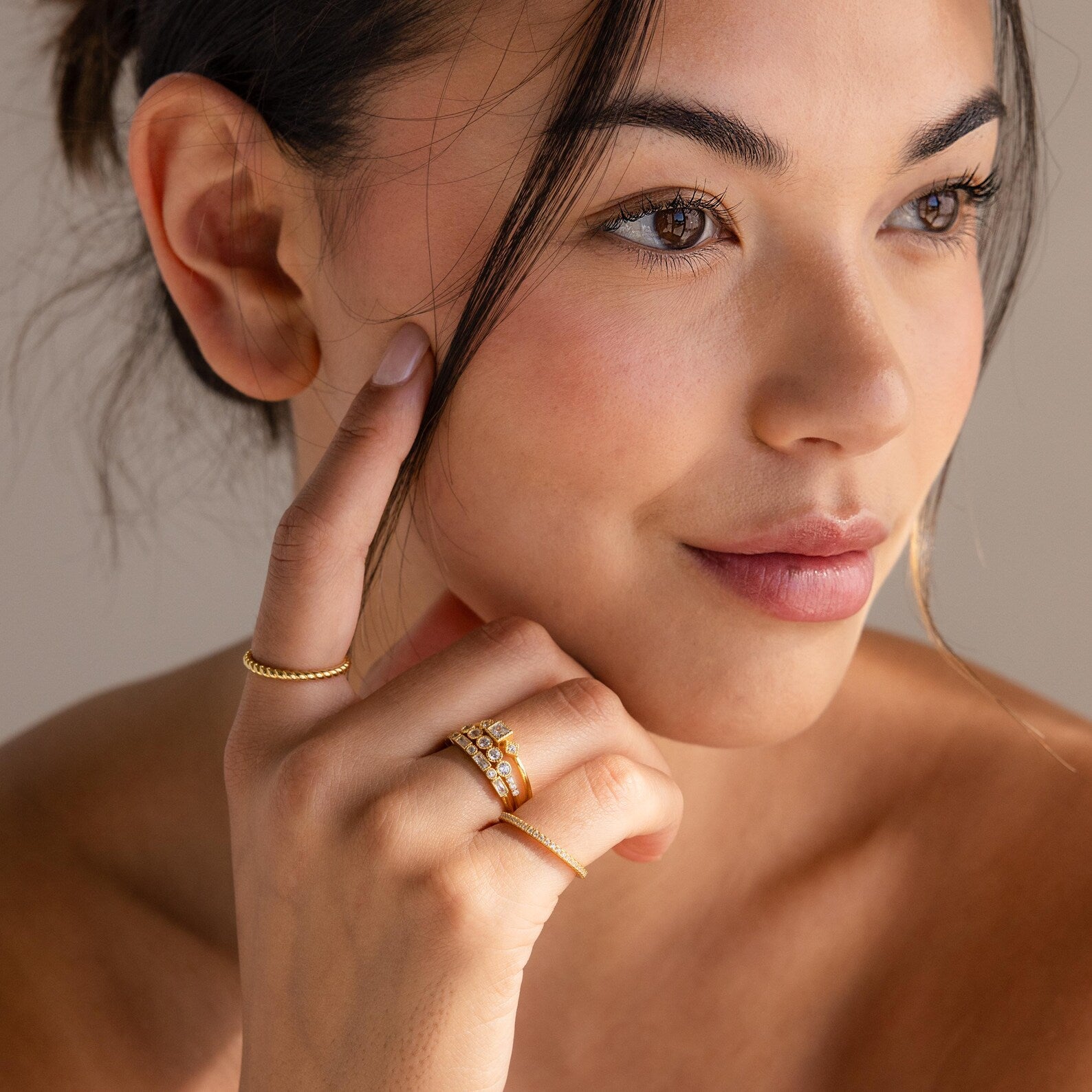 Model smiling softly while wearing multiple gold rings, including a stacked diamond Art Deco set, with her hand resting near her cheek.