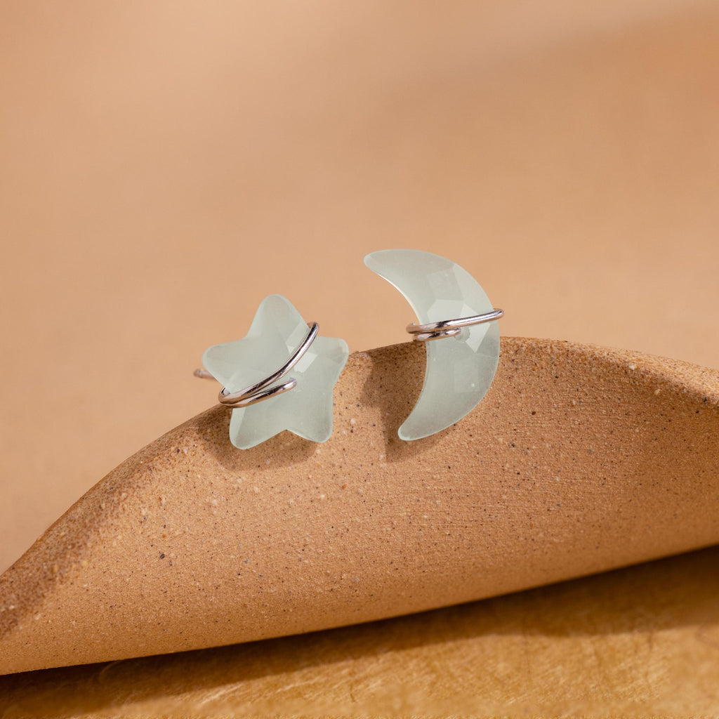The Aquamarine Moon & Star Studs, featuring one star and one crescent moon design with wire accents, are displayed on a tan surface.