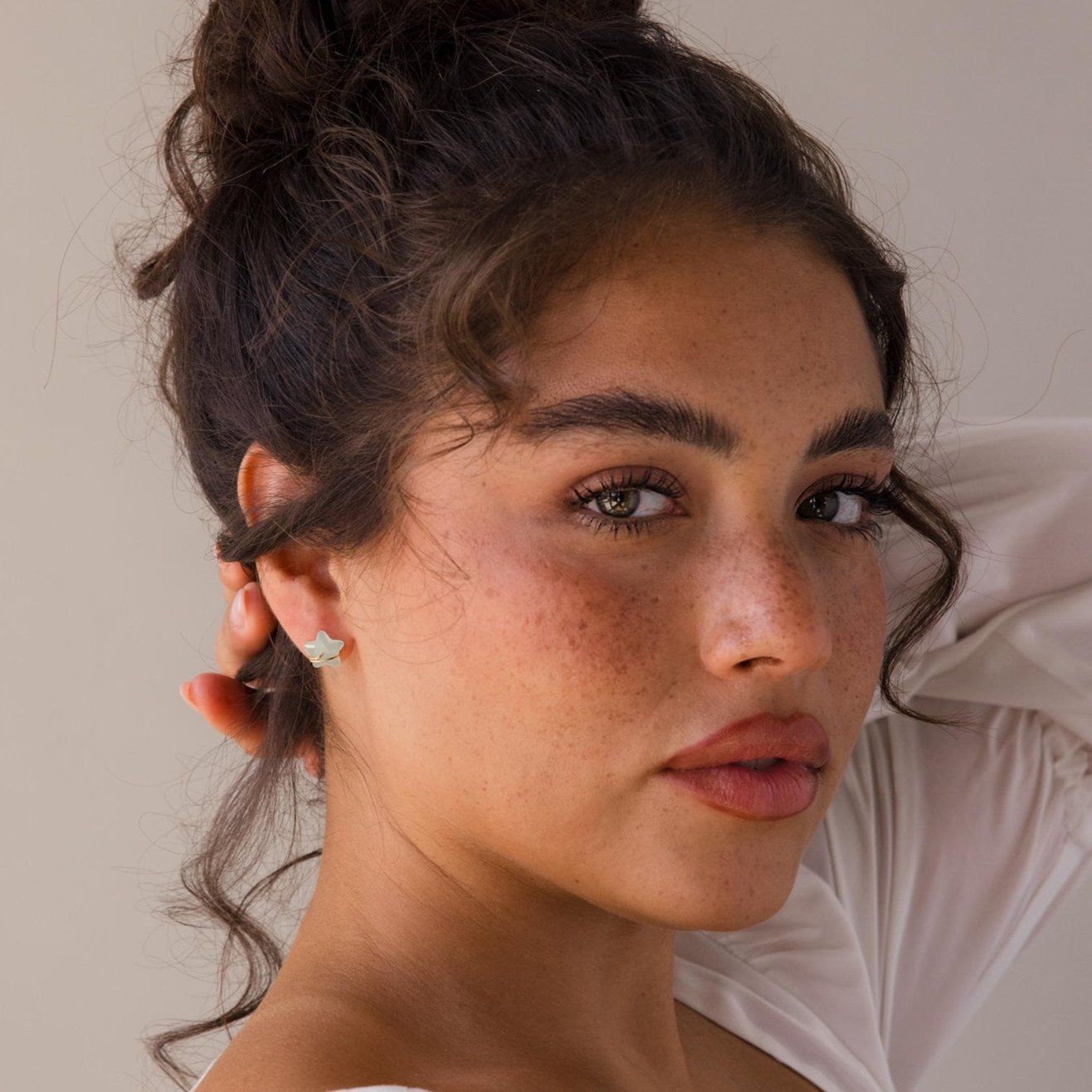 Woman with curly hair in a bun, wearing a white top and Aquamarine Moon & Star Studs, gazes at the camera.