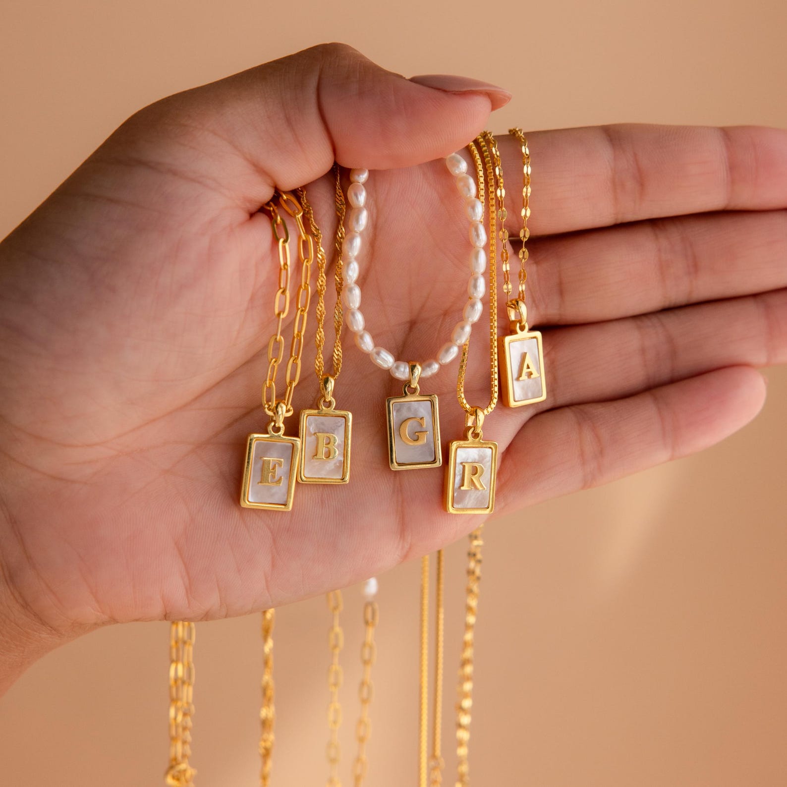 A hand displays Pearl Initial Tag Necklaces with gold rectangular pendants, each showcasing a letter—E, B, G, R, or A. This necklace makes a perfect Christmas gift and comes with a customizable chain.